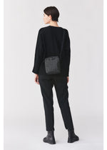 A person with short dark hair, dressed in a loose black sweater, black pants, and black boots, stands facing away. At their side hangs the BAO BAO ISSEY MIYAKE VOYAGER—a small, patterned vertical shoulder bag designed with convenient water bottle storage. The background is plain and light.