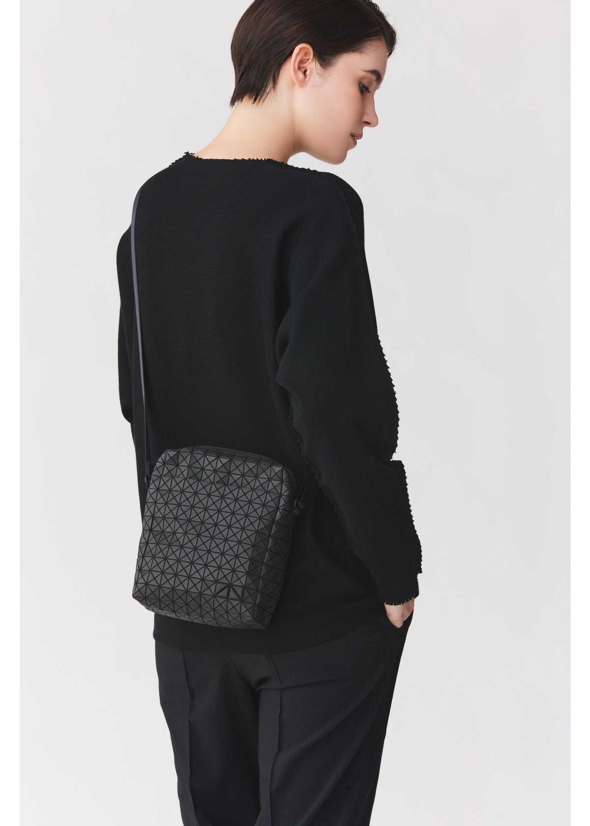 A person with short dark hair, dressed in a black sweater and pants, stands sideways with one hand in their pocket, carrying the VOYAGER by BAO BAO ISSEY MIYAKE—a sleek vertical shoulder bag adorned with a geometric pattern, perfect for travel thanks to its convenient water bottle storage. view 6 of 8