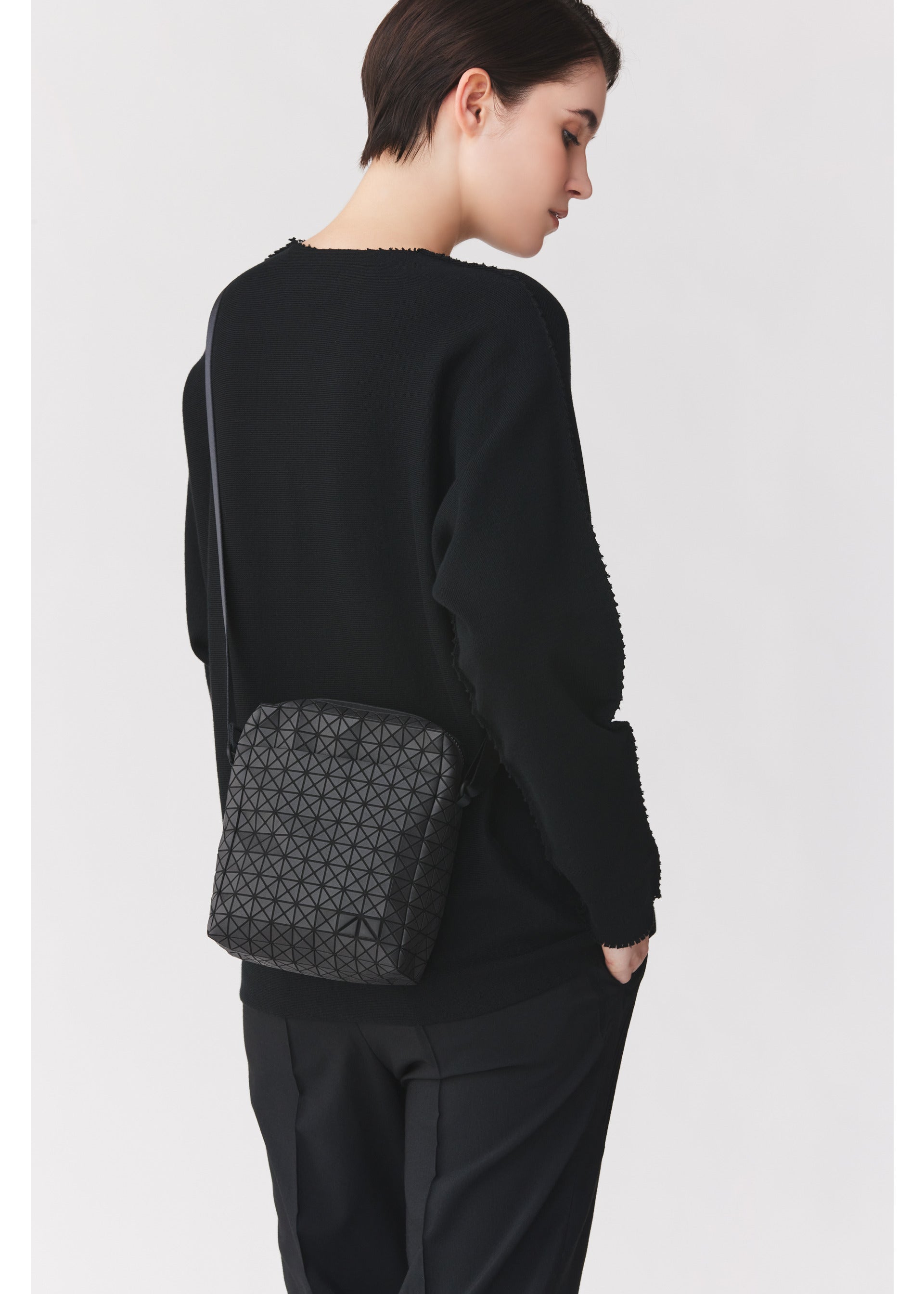 Zoomed view A person with short dark hair, dressed in a black sweater and pants, stands sideways with one hand in their pocket, carrying the VOYAGER by BAO BAO ISSEY MIYAKE—a sleek vertical shoulder bag adorned with a geometric pattern, perfect for travel thanks to its convenient water bottle storage.