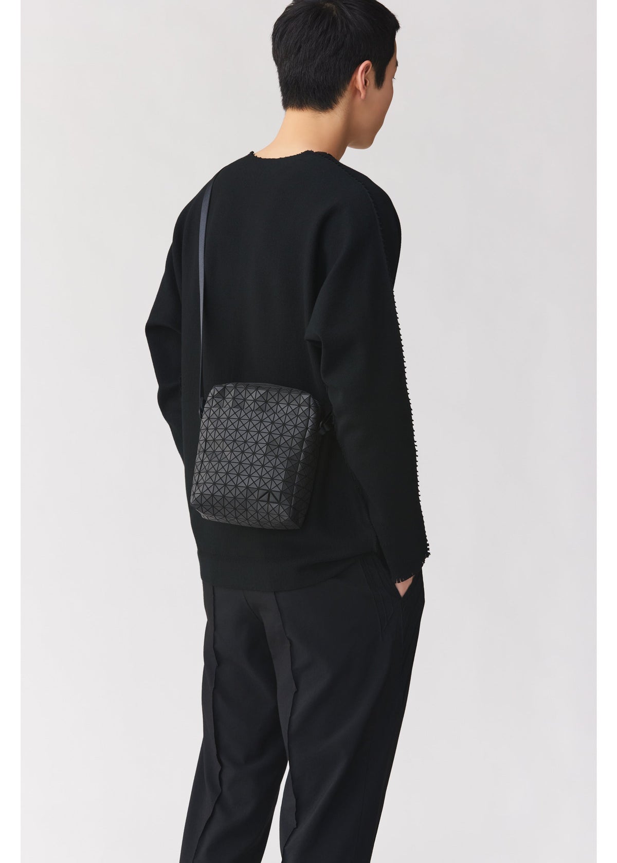 A person with short dark hair, wearing a black sweater and black pants, stands facing away from the camera with the BAO BAO ISSEY MIYAKE VOYAGER geometric-patterned black vertical shoulder bag, which features convenient water bottle storage. view 7 of 8