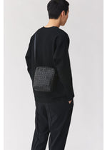 A person with short dark hair, wearing a black sweater and black pants, stands facing away from the camera with the BAO BAO ISSEY MIYAKE VOYAGER geometric-patterned black vertical shoulder bag, which features convenient water bottle storage.