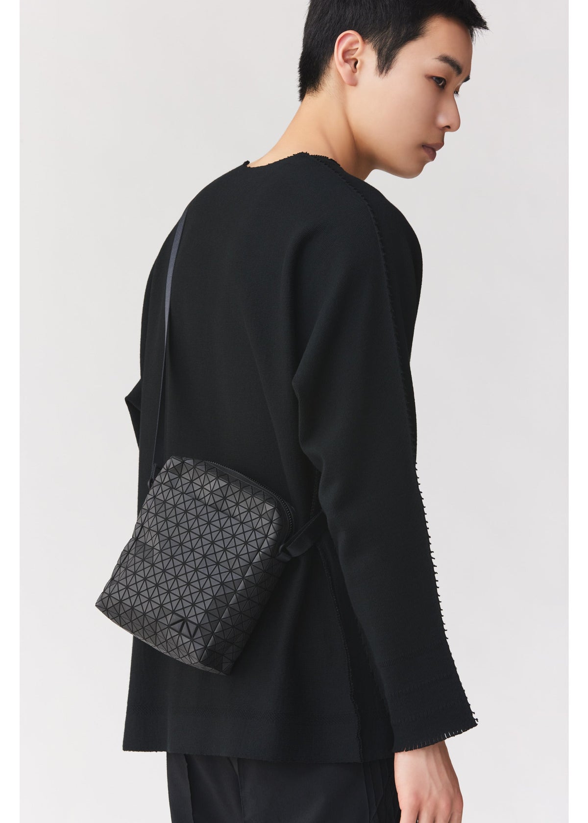 A person with short black hair wears a black long-sleeve top and carries the BAO BAO ISSEY MIYAKE VOYAGER, a textured black vertical shoulder bag with a geometric pattern and convenient water bottle storage, standing against a plain light background. view 8 of 8