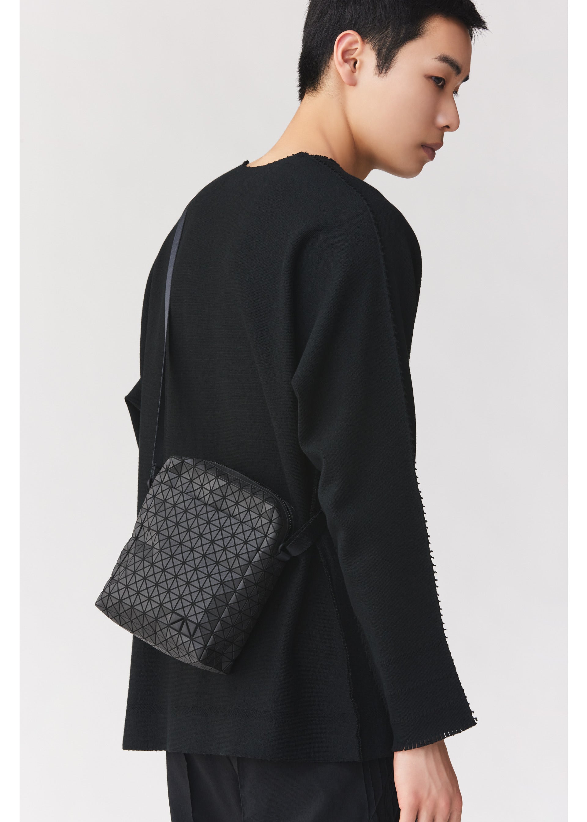 Zoomed view A person with short black hair wears a black long-sleeve top and carries the BAO BAO ISSEY MIYAKE VOYAGER, a textured black vertical shoulder bag with a geometric pattern and convenient water bottle storage, standing against a plain light background.