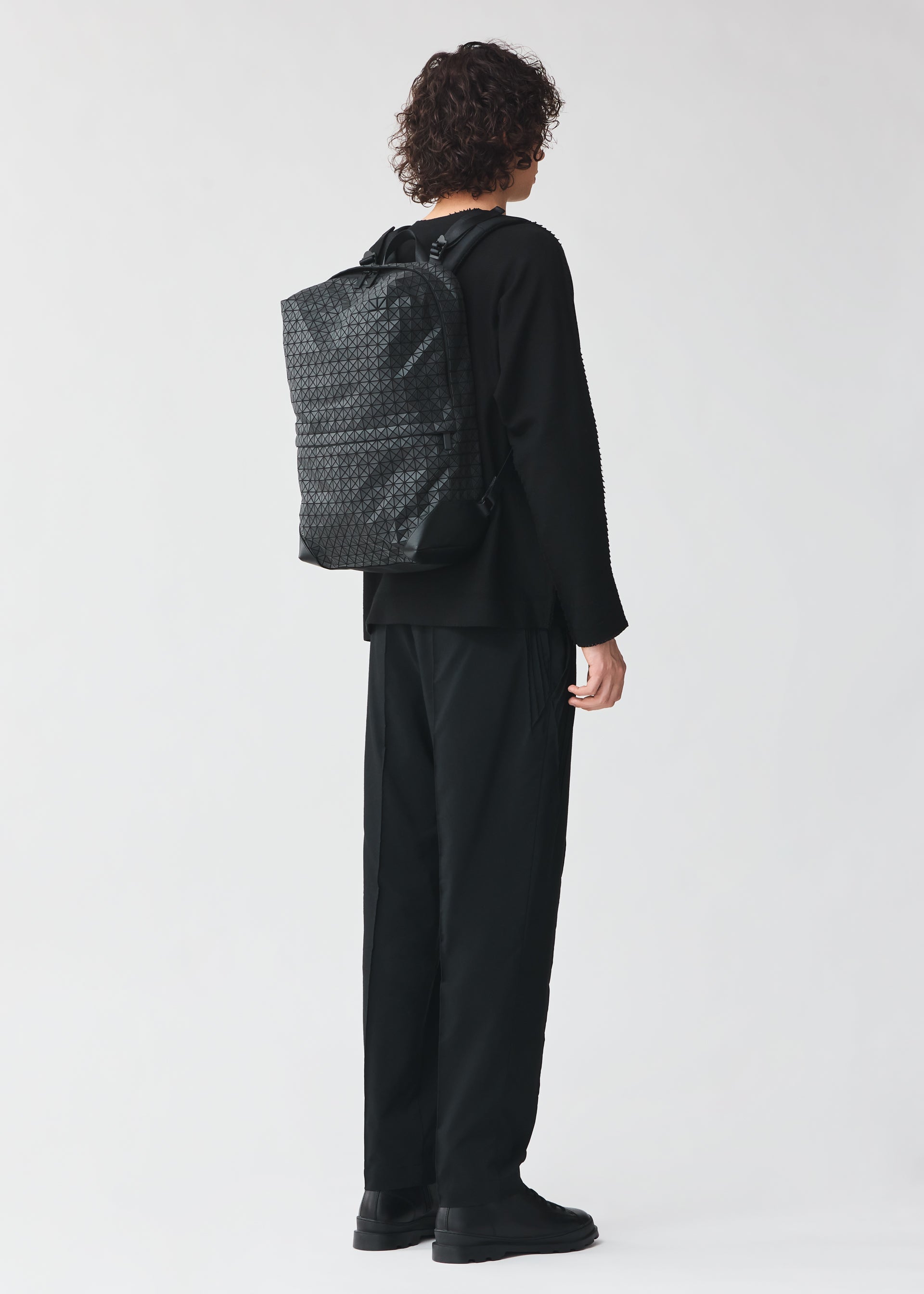 Zoomed view A person with curly hair, dressed in all black, faces away from the camera wearing a BAO BAO ISSEY MIYAKE LINER backpack with laptop storage and cowhide leather accents, set against a plain light background.