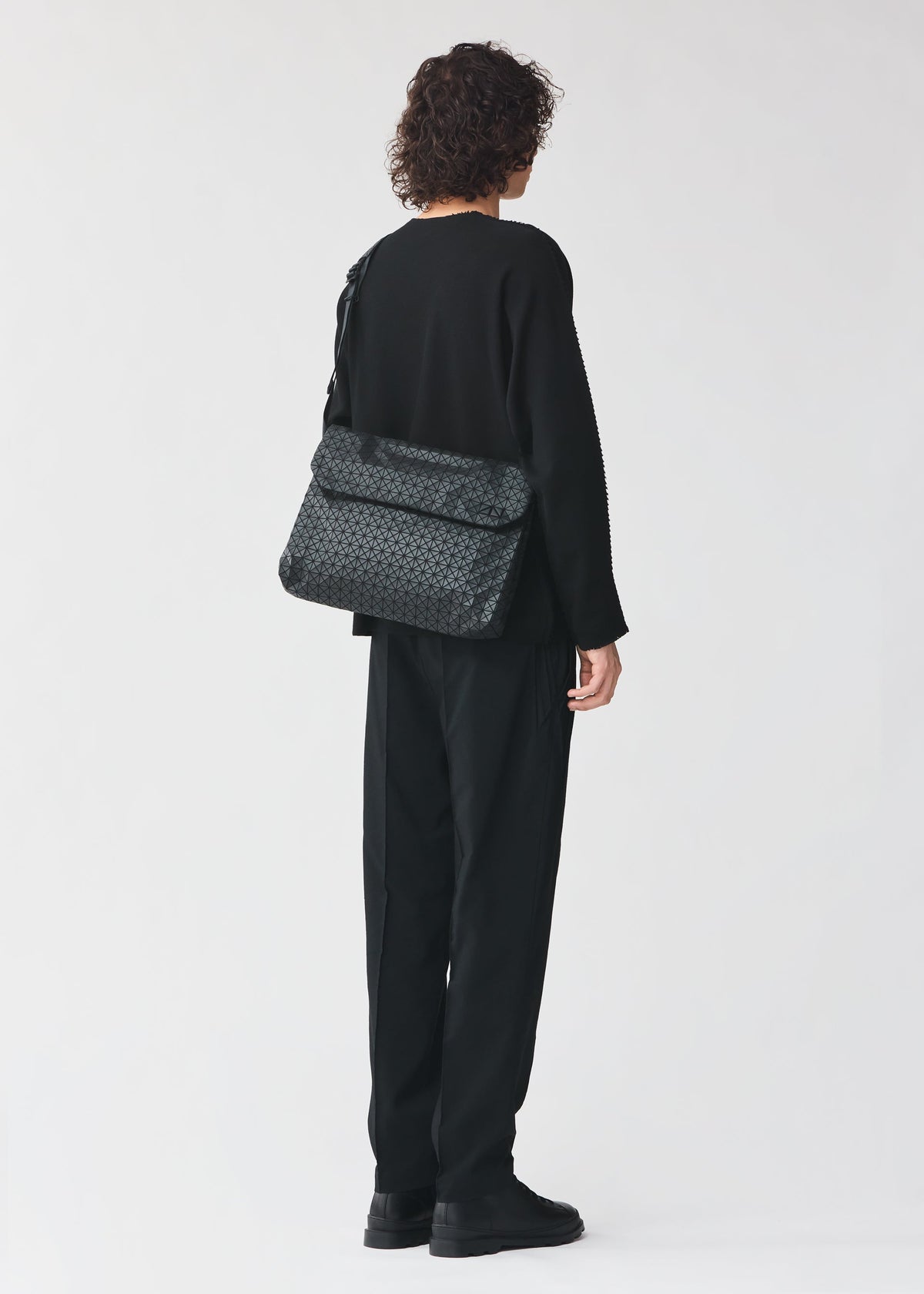 A person with curly hair, dressed in all black clothing and black shoes, stands facing away from the camera with the patterned black FROGGY shoulder bag by BAO BAO ISSEY MIYAKE—renowned for its ample storage—slung across their back against a plain light background. view 5 of 6