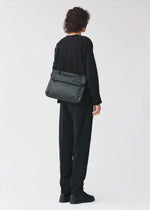 A person with curly hair, dressed in all black clothing and black shoes, stands facing away from the camera with the patterned black FROGGY shoulder bag by BAO BAO ISSEY MIYAKE—renowned for its ample storage—slung across their back against a plain light background.