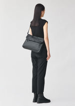 A woman with straight dark hair, dressed in a black sleeveless top, black pants, and black boots, stands facing away, carrying the BAO BAO ISSEY MIYAKE FROGGY patterned shoulder bag, renowned for its ample storage.