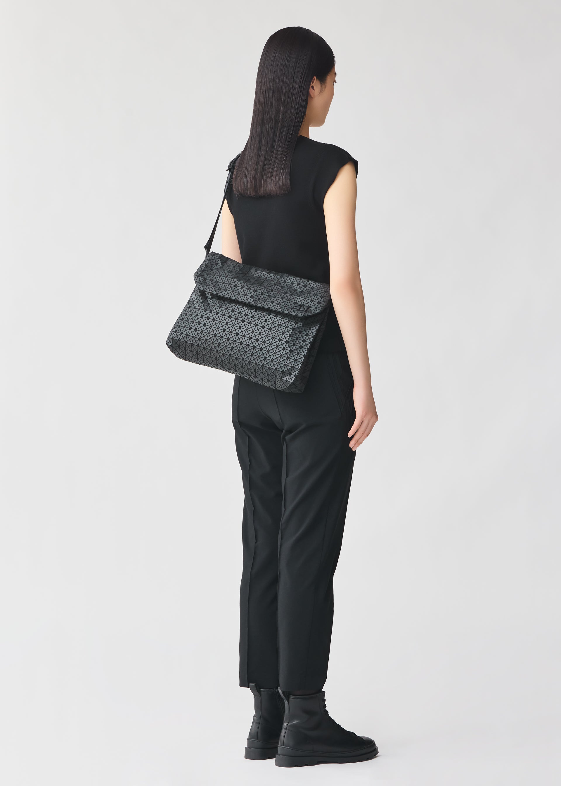 Zoomed view A woman with straight dark hair, dressed in a black sleeveless top, black pants, and black boots, stands facing away, carrying the BAO BAO ISSEY MIYAKE FROGGY patterned shoulder bag, renowned for its ample storage.
