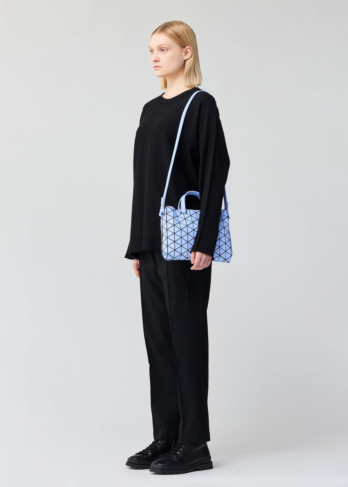 A person stands sideways against a plain light background wearing all black and carrying the TONNEAU MATTE barrel-shaped mini handbag by BAO BAO ISSEY MIYAKE, featuring a leather-like finish and geometric light blue pattern. view 6 of 6