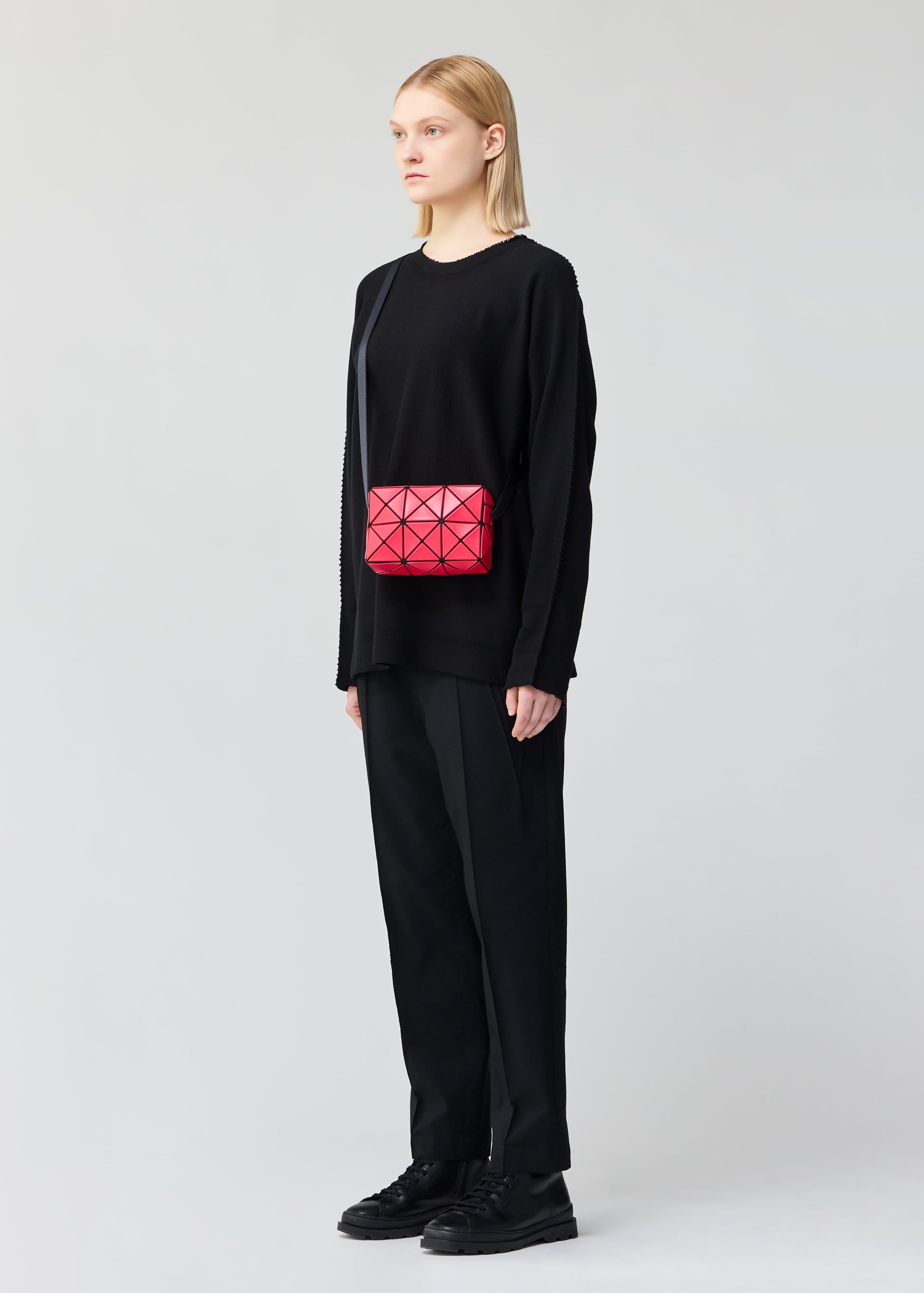Zoomed view A woman in black carries the CUBOID shoulder bag by BAO BAO ISSEY MIYAKE, featuring a pink hue and adjustable strap, accentuating her chic, minimalist style.