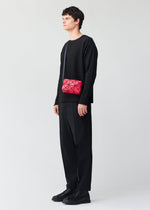 A person in black attire stands sideways against a plain light background, wearing the BAO BAO ISSEY MIYAKE CUBOID—a small red geometric-patterned shoulder bag with an adjustable strap.