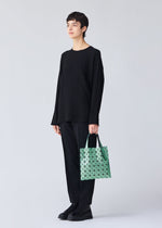 A person with short dark hair, dressed in black, stands holding the BAO BAO ISSEY MIYAKE PLATINUM shoulder bag, crafted from metallic triangular pieces, against a plain light gray background.