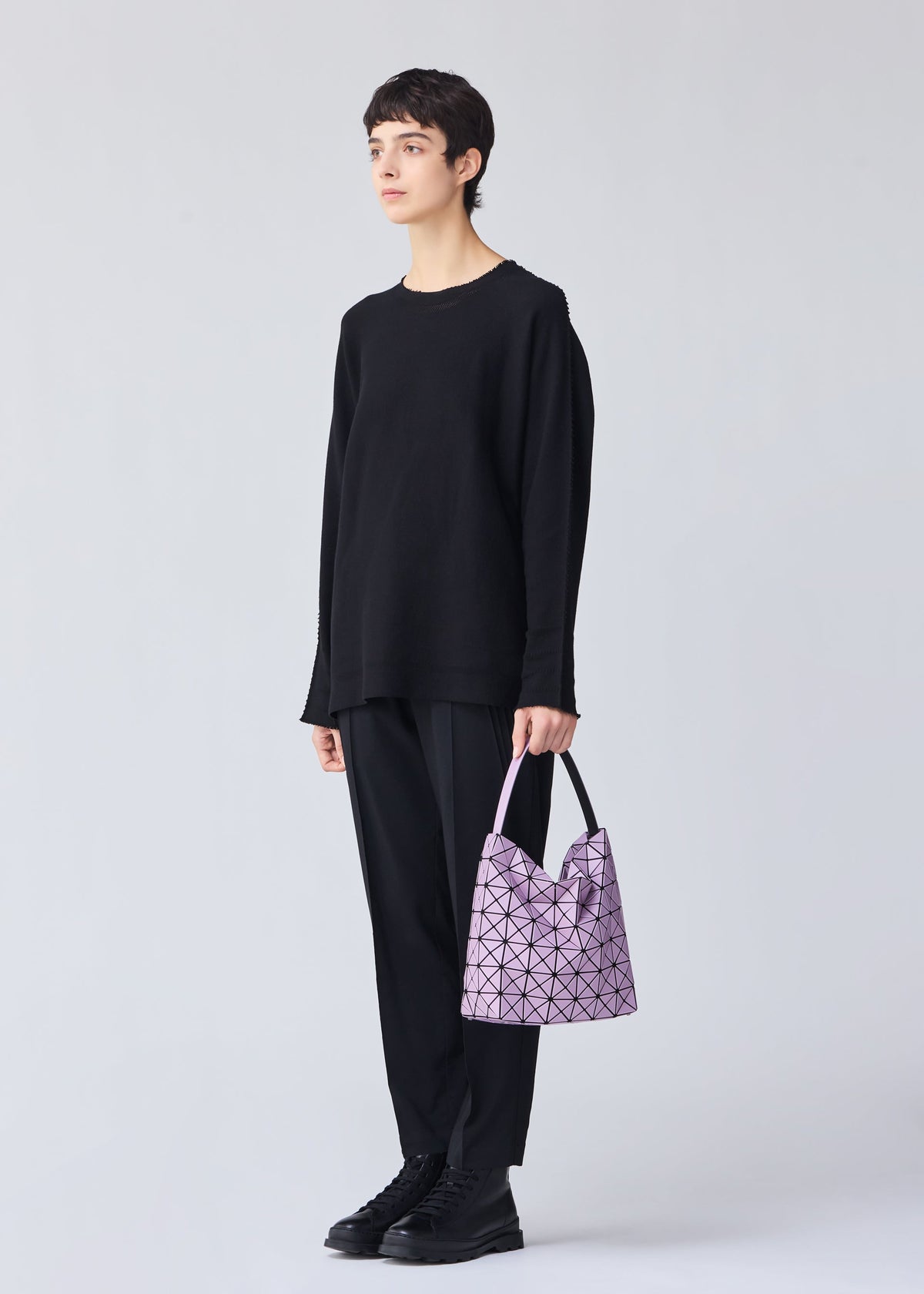 A person with short dark hair, dressed in loose black attire and boots, stands against a light background holding the VALLEY pale purple gusseted handbag by BAO BAO ISSEY MIYAKE, featuring an adjustable handle. view 6 of 6
