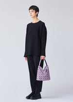 A person with short dark hair, dressed in loose black attire and boots, stands against a light background holding the VALLEY pale purple gusseted handbag by BAO BAO ISSEY MIYAKE, featuring an adjustable handle.