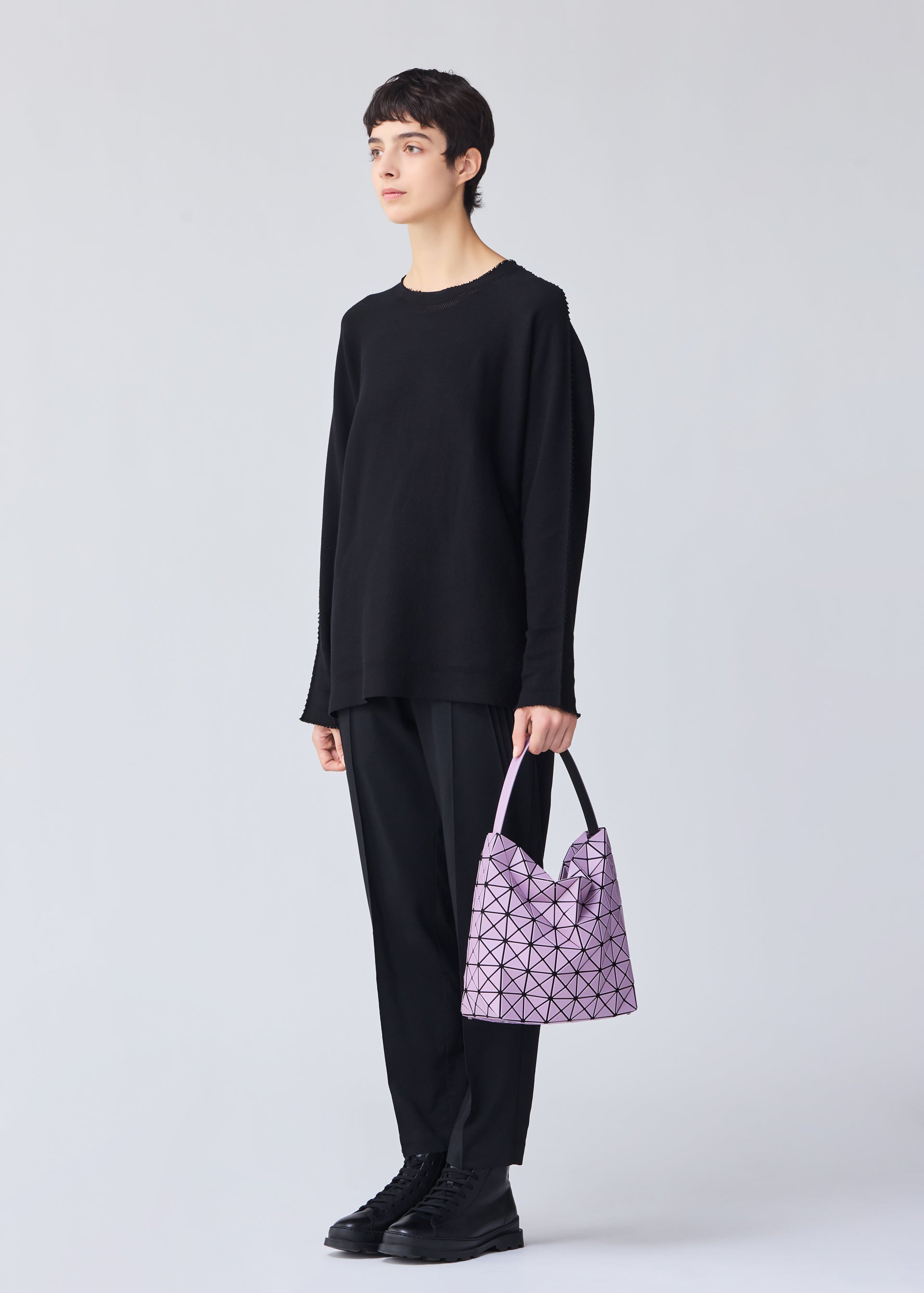 Zoomed view A person with short dark hair, dressed in loose black attire and boots, stands against a light background holding the VALLEY pale purple gusseted handbag by BAO BAO ISSEY MIYAKE, featuring an adjustable handle.