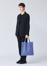 A person with short dark hair in a black suit and shoes holds a large BAO BAO ISSEY MIYAKE PRISM MATTE-2 tote featuring blue geometric patterns, perfect for carrying A4 documents, against a plain light background.