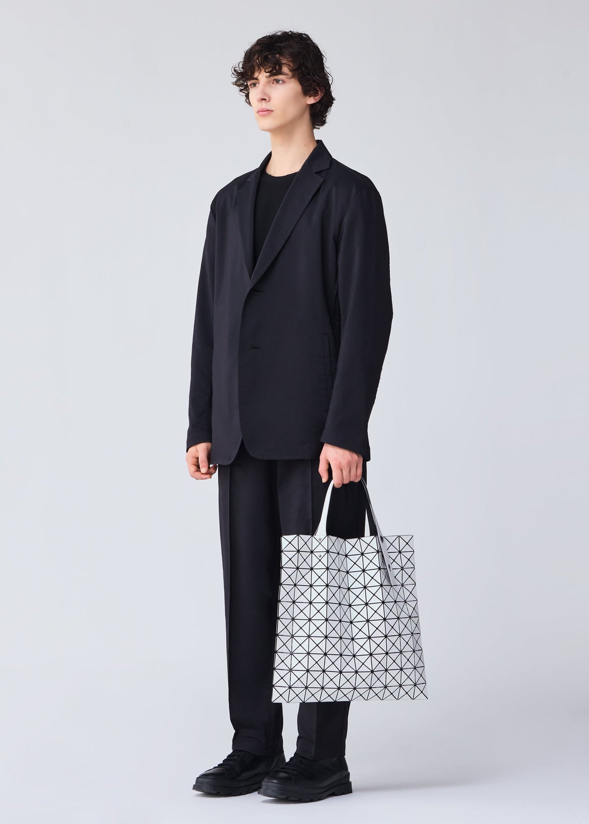 A person with short curly hair, dressed in a black suit and shoes, stands against a light background holding the BAO BAO ISSEY MIYAKE PRISM MATTE-2 tote bag, featuring a fabric grain texture and sized for A4 documents. view 8 of 8
