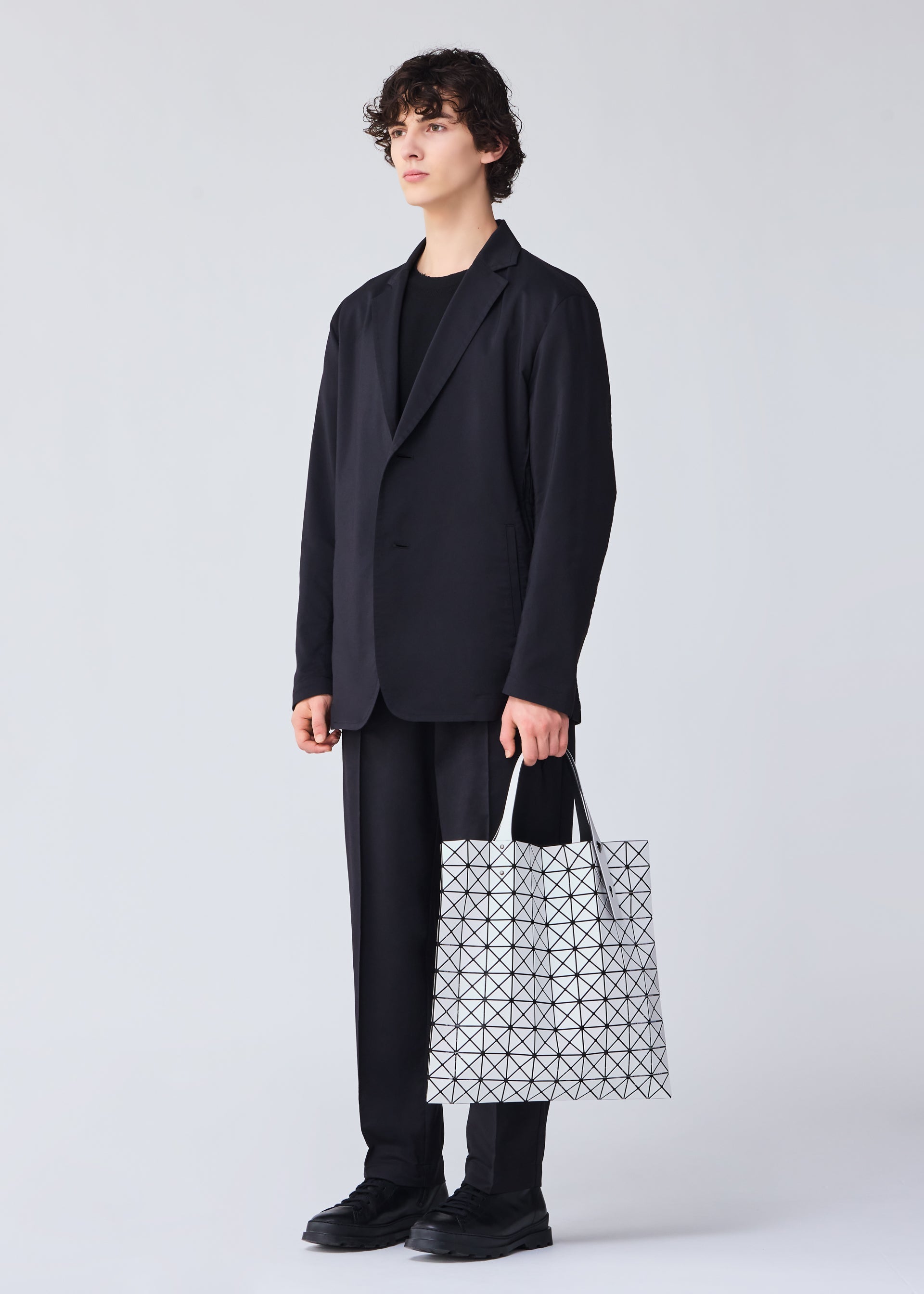 Zoomed view A person with short curly hair, dressed in a black suit and shoes, stands against a light background holding the BAO BAO ISSEY MIYAKE PRISM MATTE-2 tote bag, featuring a fabric grain texture and sized for A4 documents.