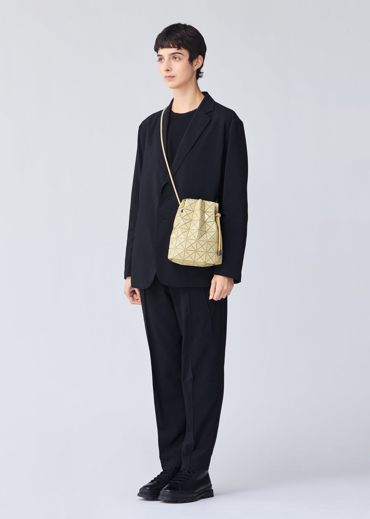 A person dressed in black stands facing forward against a plain light background, wearing the BAO BAO ISSEY MIYAKE WRING ONE-TONE, a small yellow geometric drawstring pouch, styled as a crossbody bag. view 8 of 9