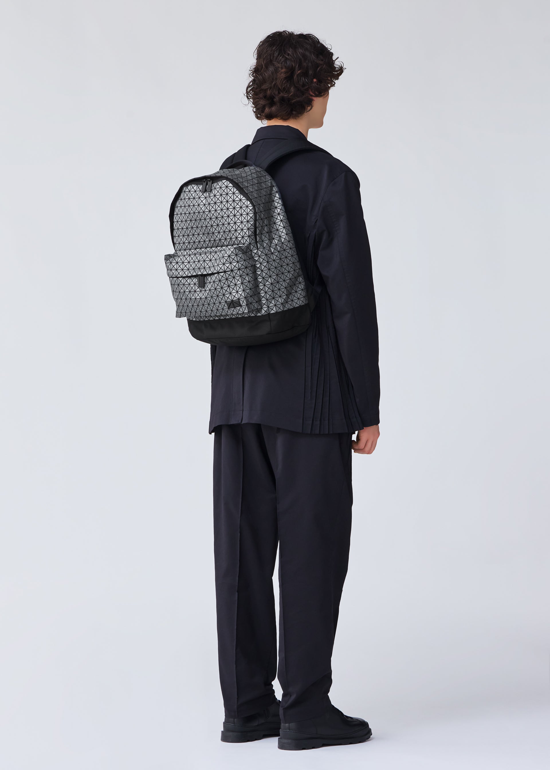Zoomed view A person with curly hair, wearing all-black clothing and shoes, faces away from the camera with a grey DAYPACK REFLECTOR by BAO BAO ISSEY MIYAKE on their back. The background is plain and light-colored.