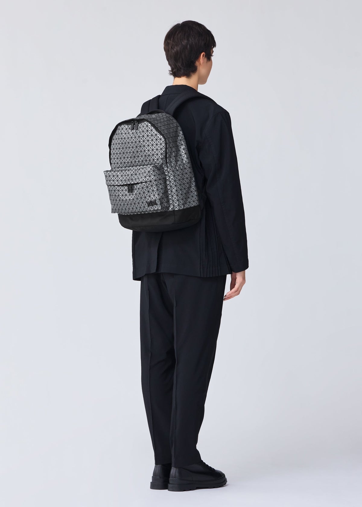 A person with short dark hair, dressed in all-black, stands facing away from the camera, wearing the BAO BAO ISSEY MIYAKE DAYPACK REFLECTOR— a unisex backpack with gray patterns and black accents—against a plain light background. view 6 of 7
