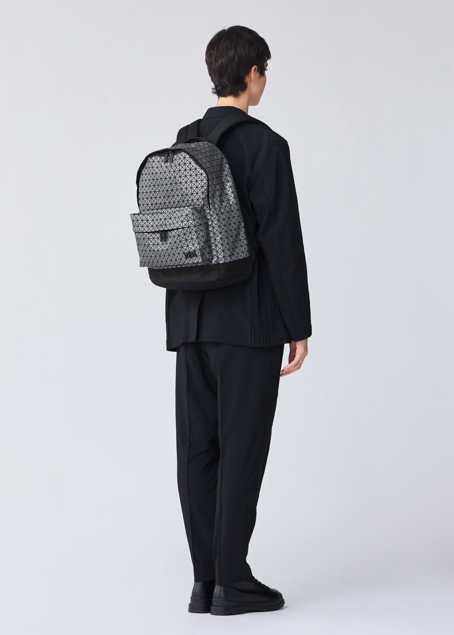 Zoomed view A person with short dark hair, dressed in all-black, stands facing away from the camera, wearing the BAO BAO ISSEY MIYAKE DAYPACK REFLECTOR— a unisex backpack with gray patterns and black accents—against a plain light background.