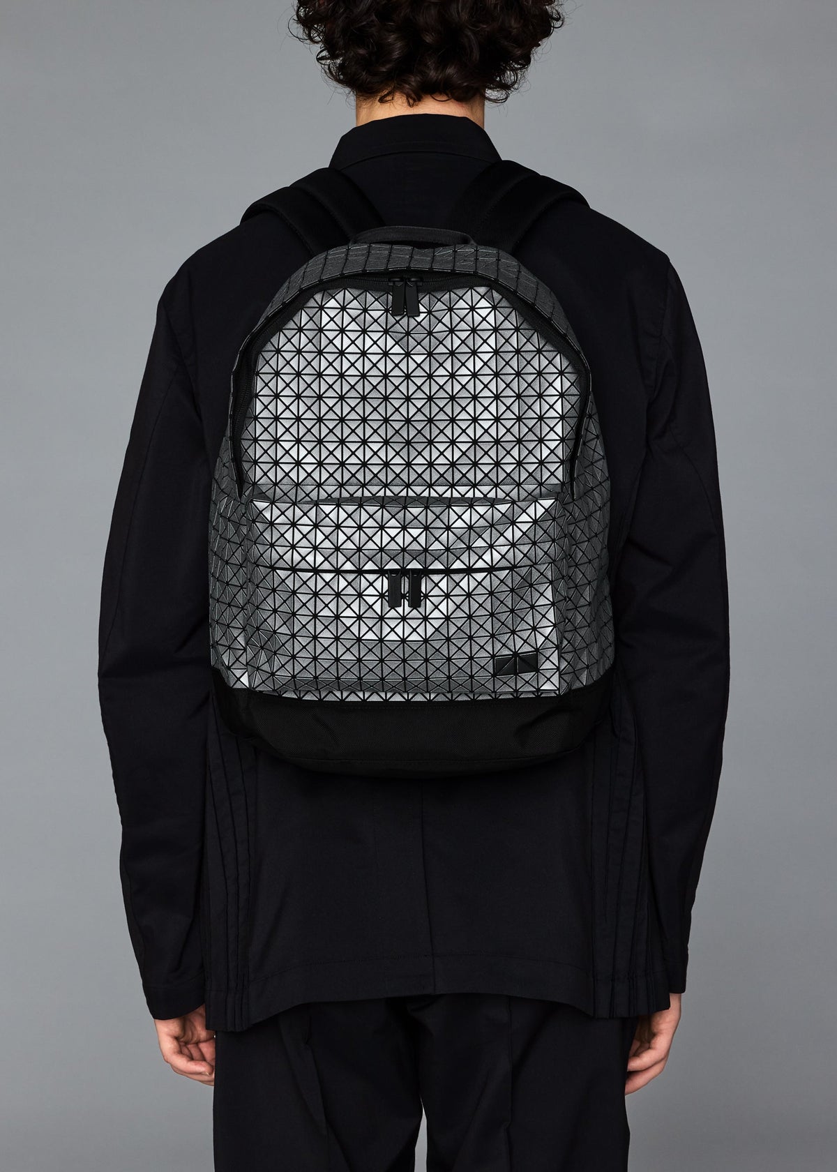A person with curly hair in black clothing is seen from the back wearing the BAO BAO ISSEY MIYAKE DAYPACK REFLECTOR, a unisex backpack with a geometric triangular pattern. The background is plain gray. view 7 of 7