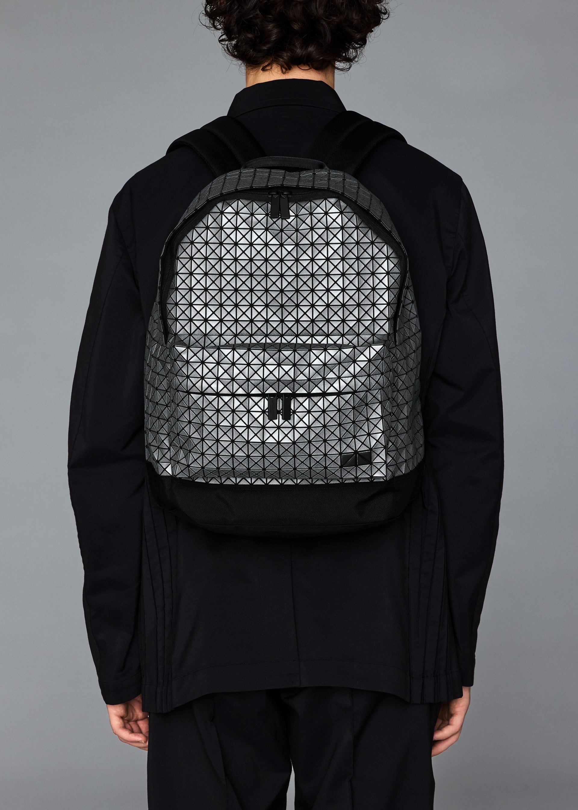 Zoomed view A person with curly hair in black clothing is seen from the back wearing the BAO BAO ISSEY MIYAKE DAYPACK REFLECTOR, a unisex backpack with a geometric triangular pattern. The background is plain gray.