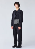 A person with curly hair wears a loose black suit, black shirt, and black shoes against a light background, carrying the BAO BAO ISSEY MIYAKE VOYAGER REFLECTOR—a stylish gray metallic tote—across their body.