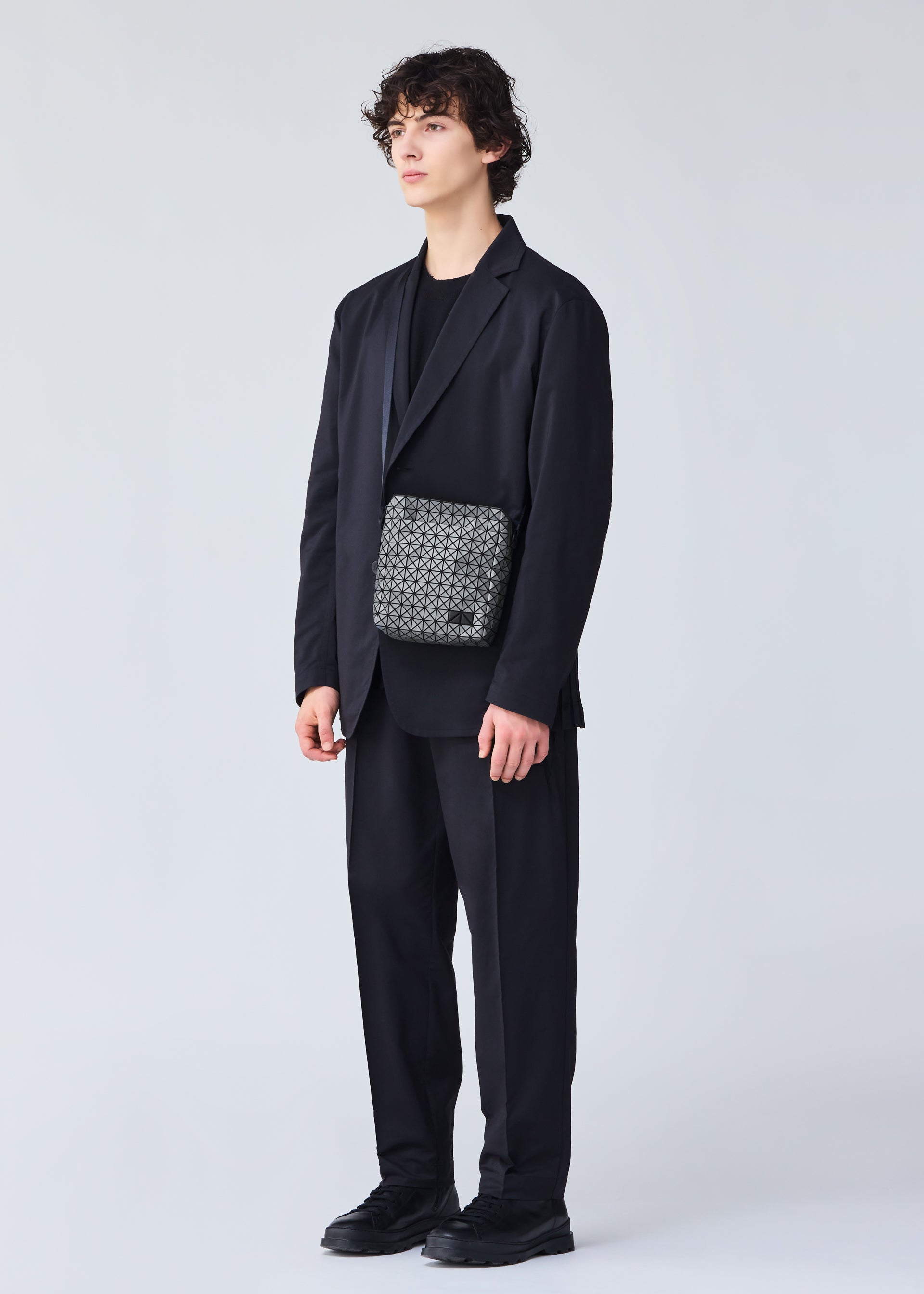 Zoomed view A person with curly hair wears a loose black suit, black shirt, and black shoes against a light background, carrying the BAO BAO ISSEY MIYAKE VOYAGER REFLECTOR—a stylish gray metallic tote—across their body.