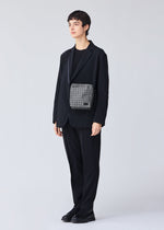 A person with short dark hair stands against a light background, wearing a loose black suit and shoes, accessorized with the BAO BAO ISSEY MIYAKE VOYAGER REFLECTOR tote bag worn crossbody.