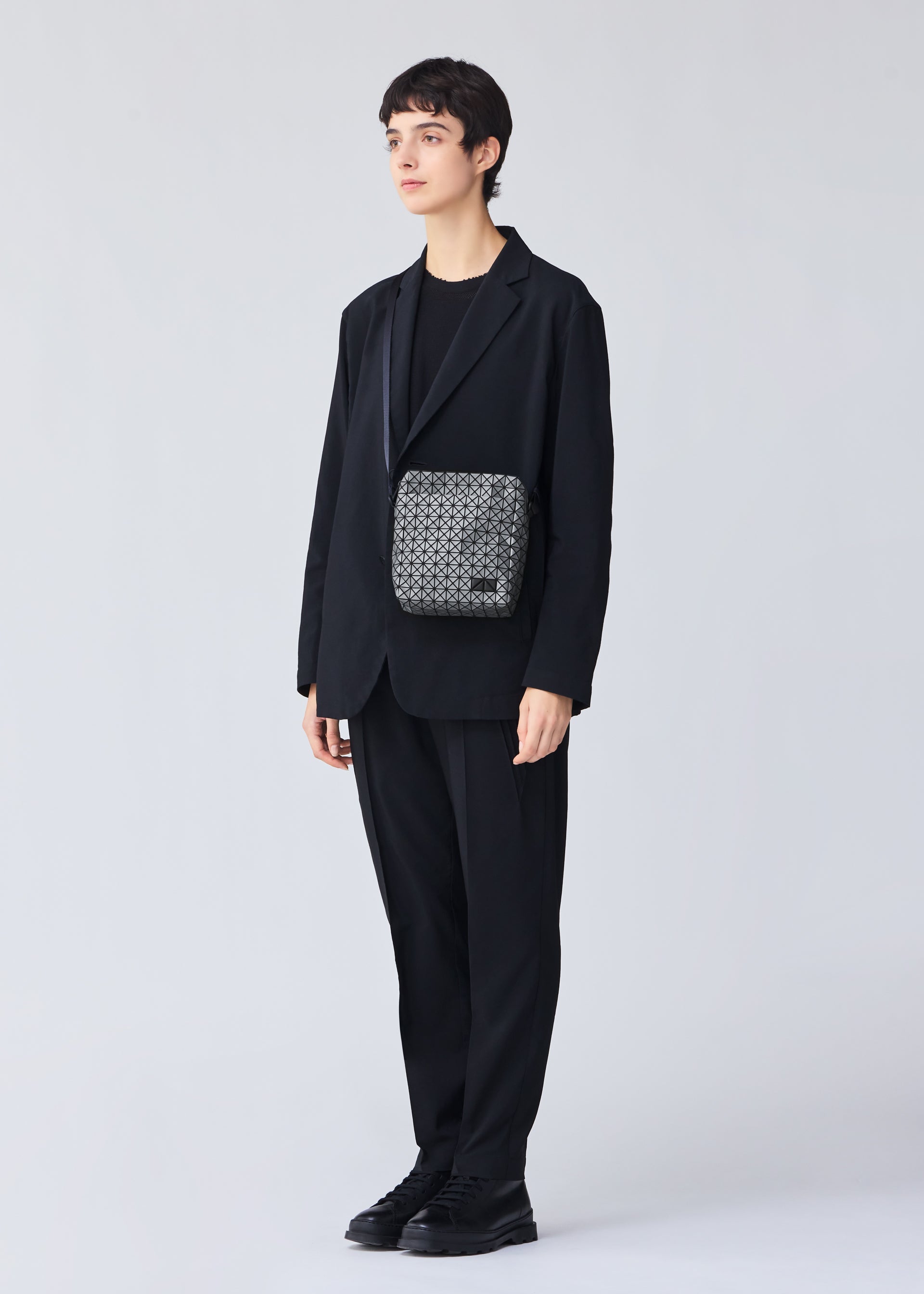 Zoomed view A person with short dark hair stands against a light background, wearing a loose black suit and shoes, accessorized with the BAO BAO ISSEY MIYAKE VOYAGER REFLECTOR tote bag worn crossbody.