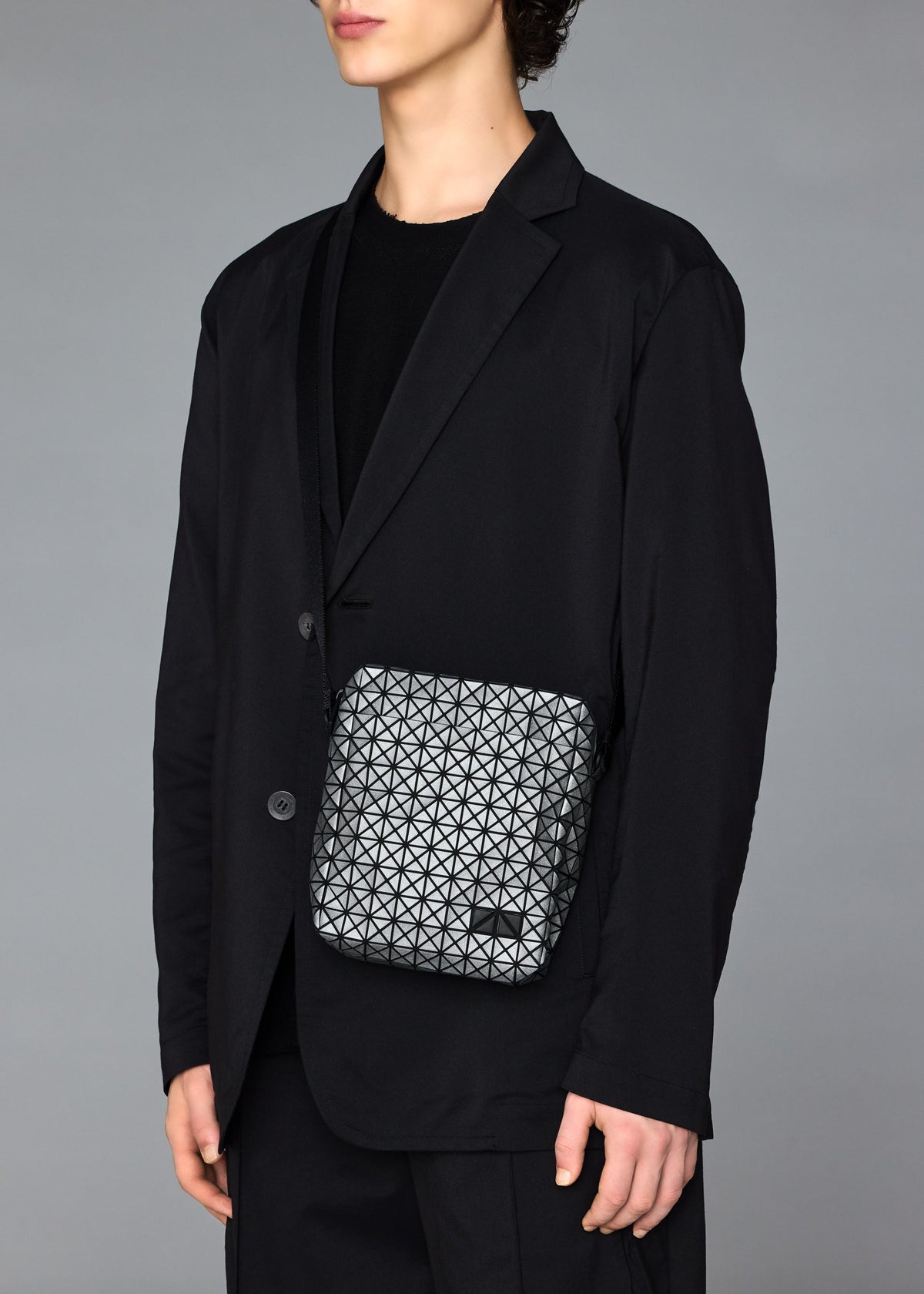 A person in a black suit and shirt holds the BAO BAO ISSEY MIYAKE VOYAGER REFLECTOR, a small geometric-patterned tote bag, against a plain gray background. Their face is partially out of frame. view 7 of 7