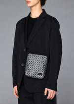 A person in a black suit and shirt holds the BAO BAO ISSEY MIYAKE VOYAGER REFLECTOR, a small geometric-patterned tote bag, against a plain gray background. Their face is partially out of frame.