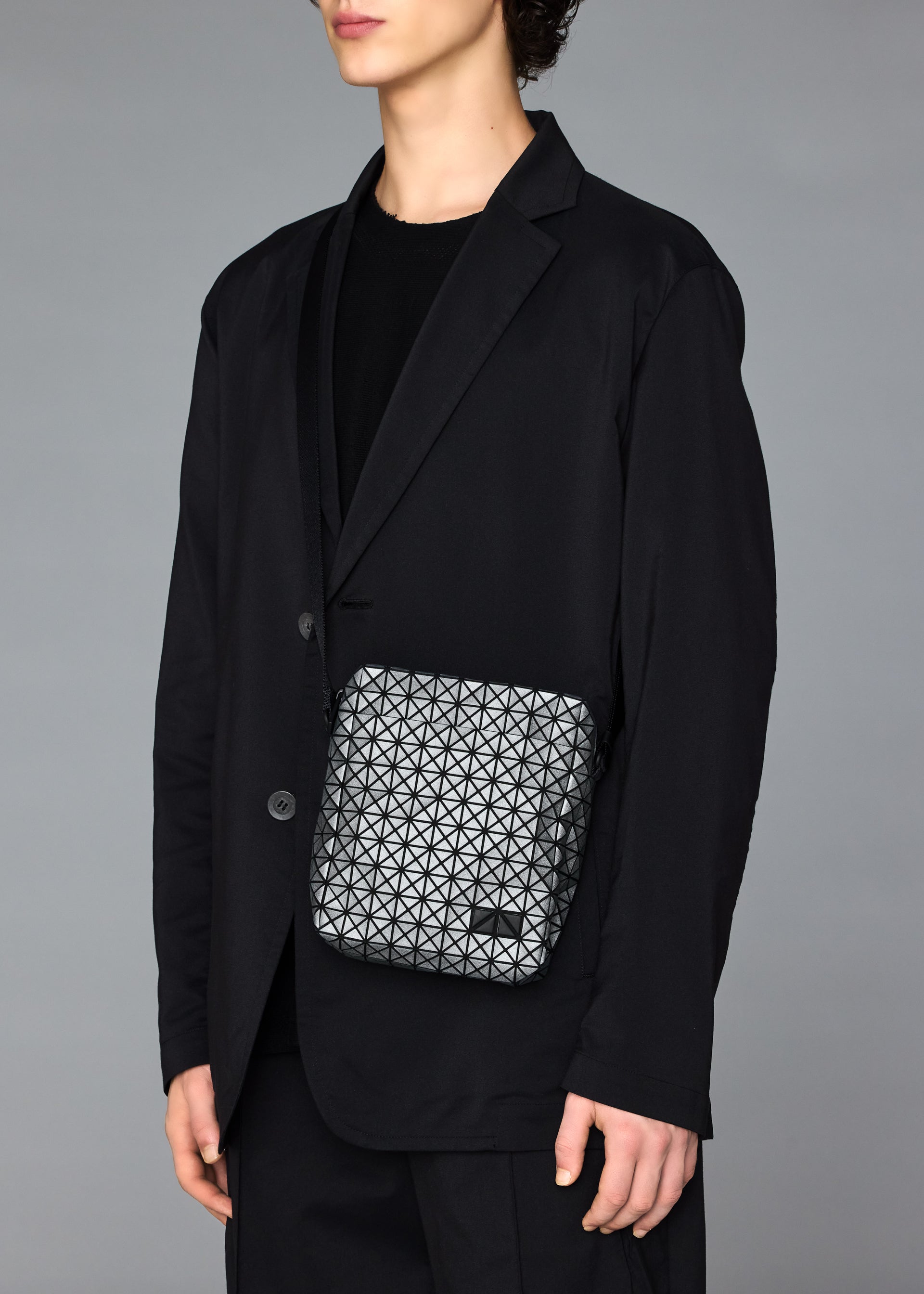 Zoomed view A person in a black suit and shirt holds the BAO BAO ISSEY MIYAKE VOYAGER REFLECTOR, a small geometric-patterned tote bag, against a plain gray background. Their face is partially out of frame.