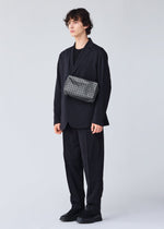 A person with curly hair in a loose black suit and shoes stands against a plain light background, wearing the BAO BAO ISSEY MIYAKE SADDLE BAG REFLECTOR—a patterned black-and-white reflective shoulder bag with an inner zipper pocket.