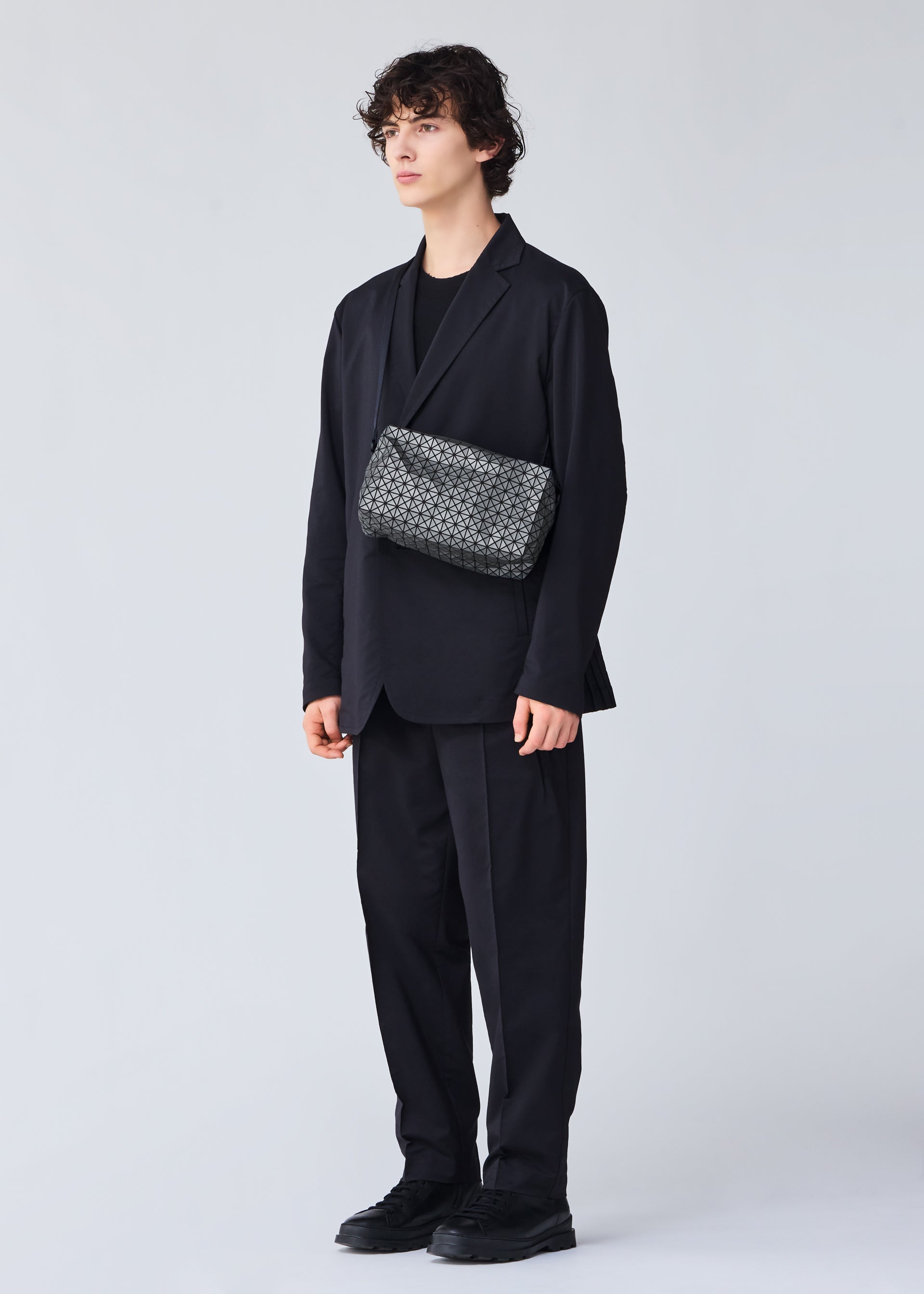 Zoomed view A person with curly hair in a loose black suit and shoes stands against a plain light background, wearing the BAO BAO ISSEY MIYAKE SADDLE BAG REFLECTOR—a patterned black-and-white reflective shoulder bag with an inner zipper pocket.