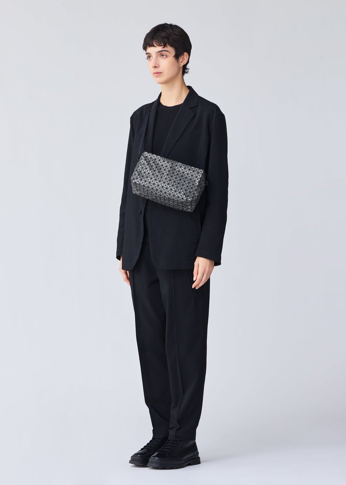 A person with short dark hair stands against a light gray background, dressed in a loose black suit and shoes, holding the BAO BAO ISSEY MIYAKE SADDLE BAG REFLECTOR featuring a subtle black and gray pattern. view 7 of 8