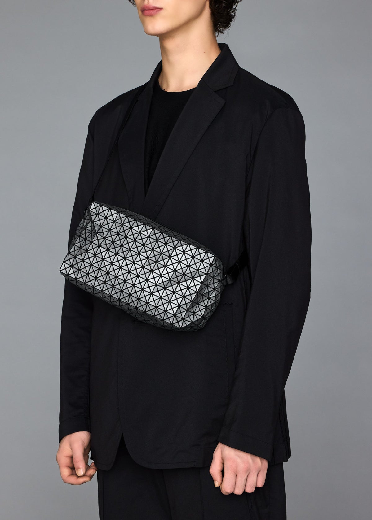 A person in a black suit and shirt stands against a gray background, carrying the BAO BAO ISSEY MIYAKE SADDLE BAG REFLECTOR—a geometric-patterned metallic bag—across their chest, with their head slightly turned and face partly out of frame. view 8 of 8
