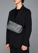 A person in a black suit and shirt stands against a gray background, carrying the BAO BAO ISSEY MIYAKE SADDLE BAG REFLECTOR—a geometric-patterned metallic bag—across their chest, with their head slightly turned and face partly out of frame.