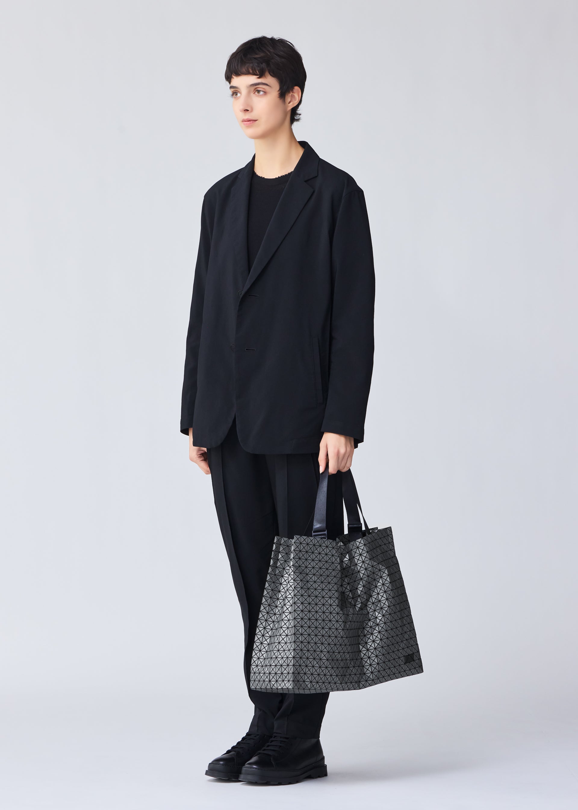Zoomed view A person with short dark hair, in a loose black suit and shoes, holds the BAO BAO ISSEY MIYAKE CART REFLECTOR metallic tote with adjustable handles against a plain light background.
