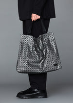 A person dressed in black holds the BAO BAO ISSEY MIYAKE CART REFLECTOR tote, featuring geometric silver panels. Only their lower body—black pants and shoes—is shown against a gray background.