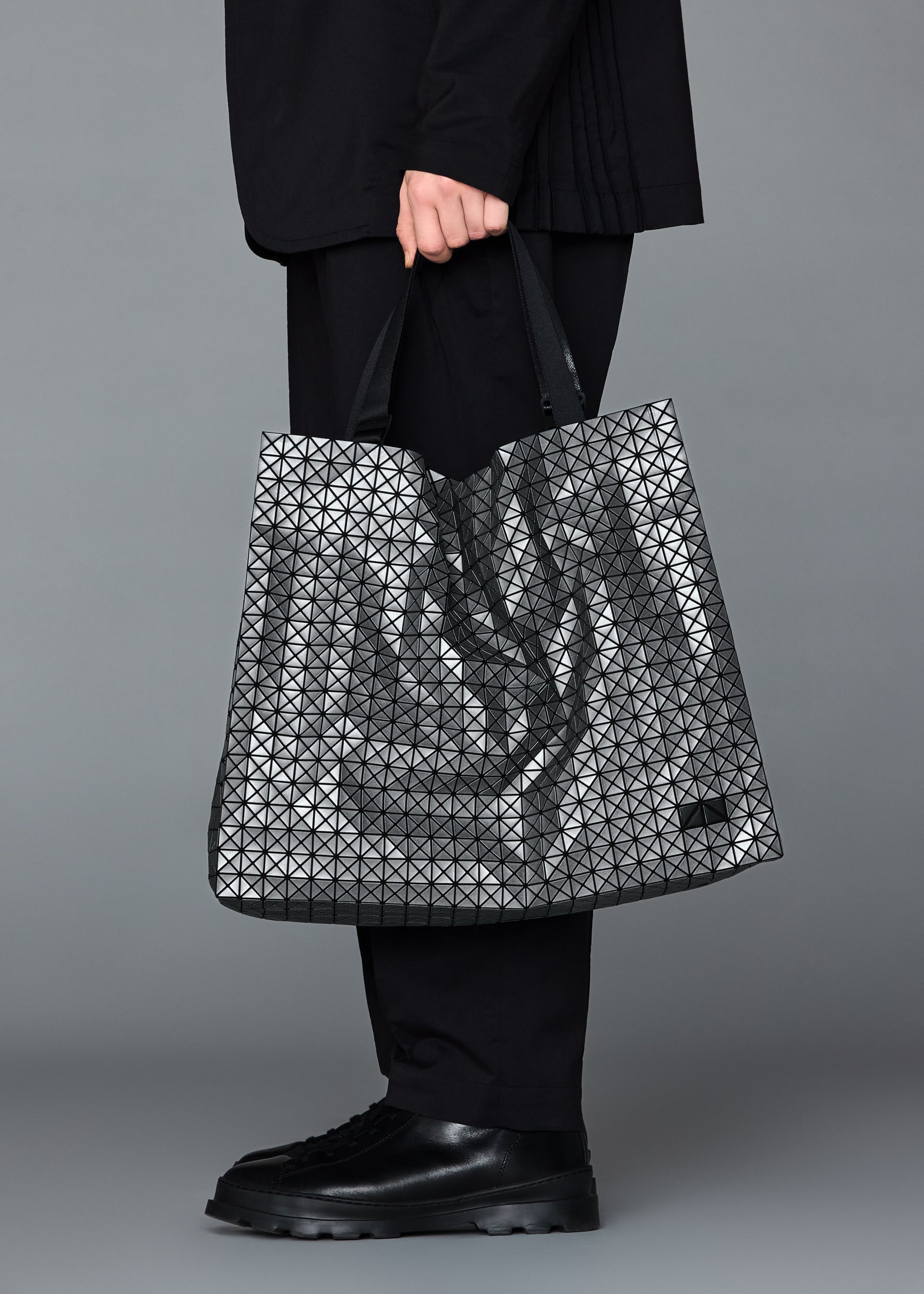 Zoomed view A person dressed in black holds the BAO BAO ISSEY MIYAKE CART REFLECTOR tote, featuring geometric silver panels. Only their lower body—black pants and shoes—is shown against a gray background.