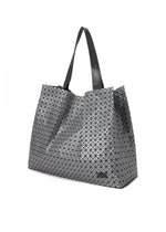 The CART REFLECTOR by BAO BAO ISSEY MIYAKE is a large metallic tote featuring a geometric triangular pattern, black adjustable handles, and a slightly glossy finish, shown against a white background.