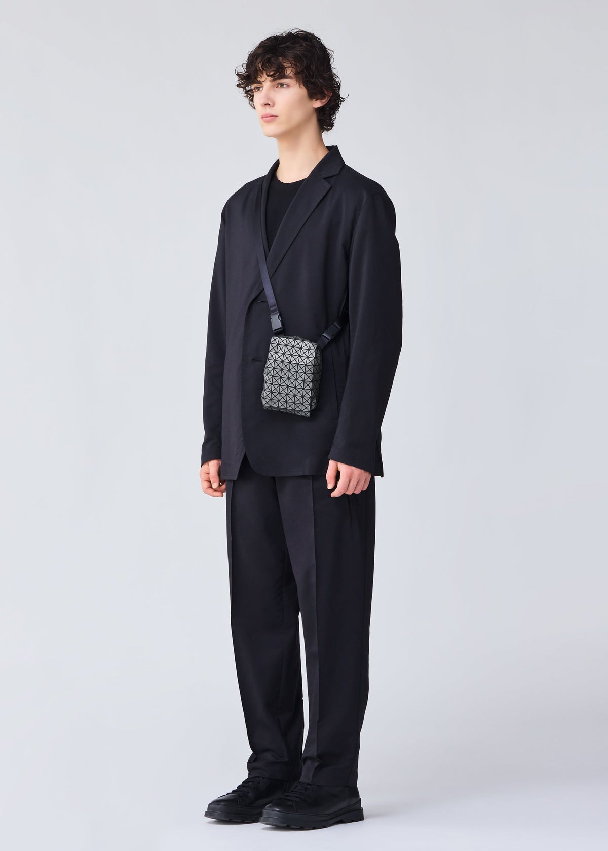 A person with short curly hair wears a loose black suit, tee, and shoes against a light gray background. They carry the BEETLE REFLECTOR crossbody bag by BAO BAO ISSEY MIYAKE, featuring a small patterned design with a shoulder strap. view 6 of 8