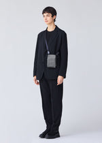 A person with short dark hair in a black suit, shirt, and shoes stands against a light gray background, carrying the BAO BAO ISSEY MIYAKE BEETLE REFLECTOR crossbody bag over one shoulder.