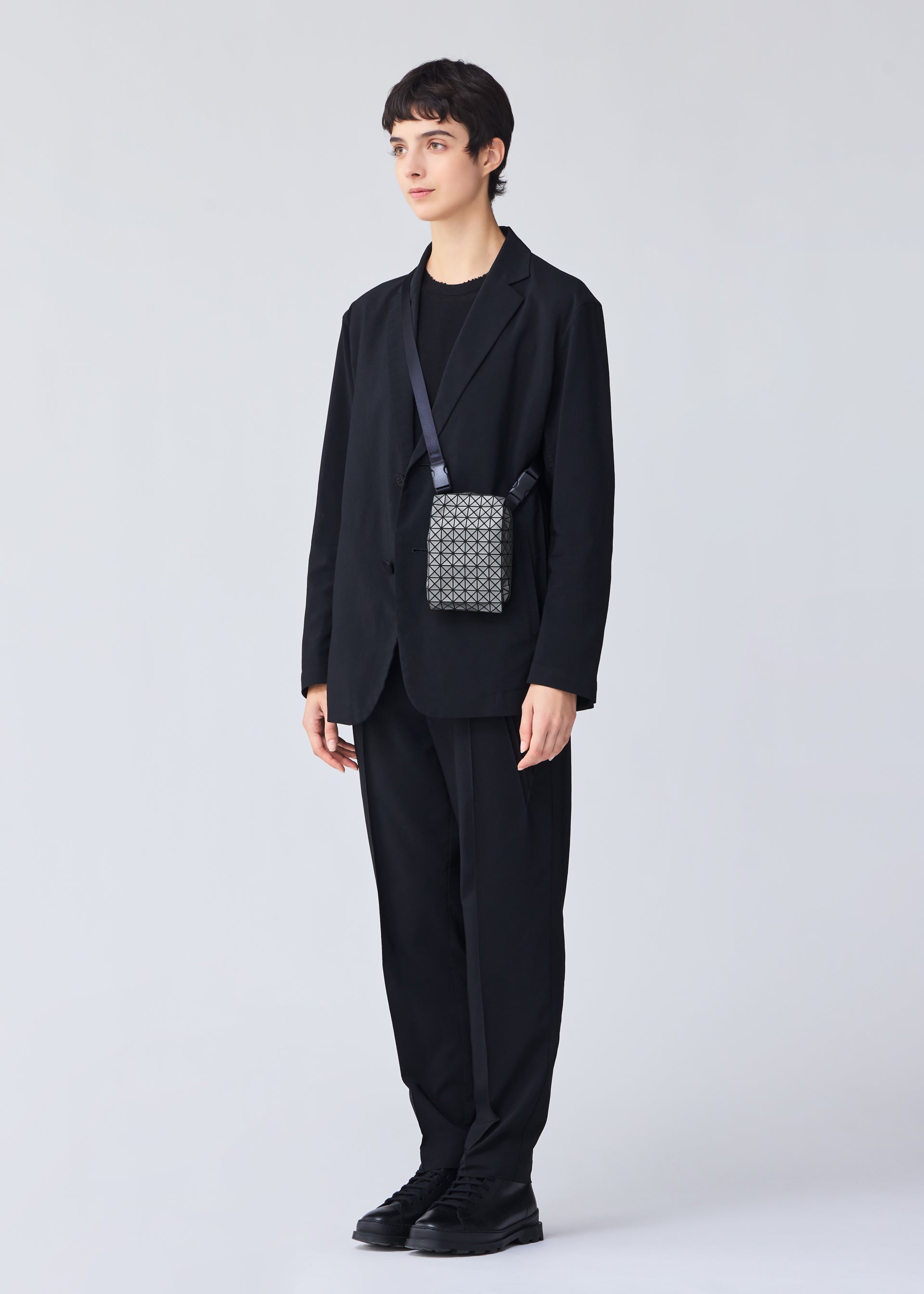Zoomed view A person with short dark hair in a black suit, shirt, and shoes stands against a light gray background, carrying the BAO BAO ISSEY MIYAKE BEETLE REFLECTOR crossbody bag over one shoulder.