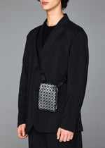 A person in a black suit and shirt stands against a plain gray background, carrying the BAO BAO ISSEY MIYAKE BEETLE REFLECTOR crossbody bag with a geometric reflective design. Their face is partially out of frame.