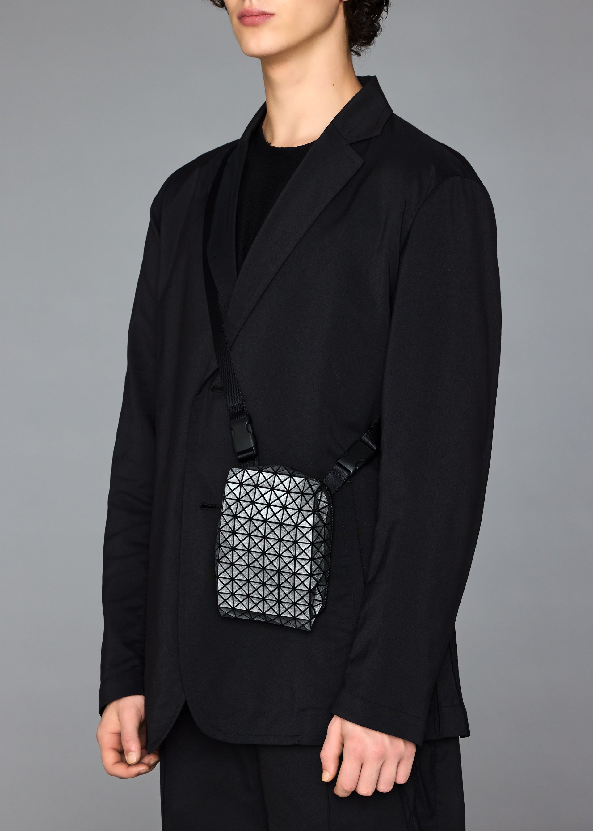 Zoomed view A person in a black suit and shirt stands against a plain gray background, carrying the BAO BAO ISSEY MIYAKE BEETLE REFLECTOR crossbody bag with a geometric reflective design. Their face is partially out of frame.
