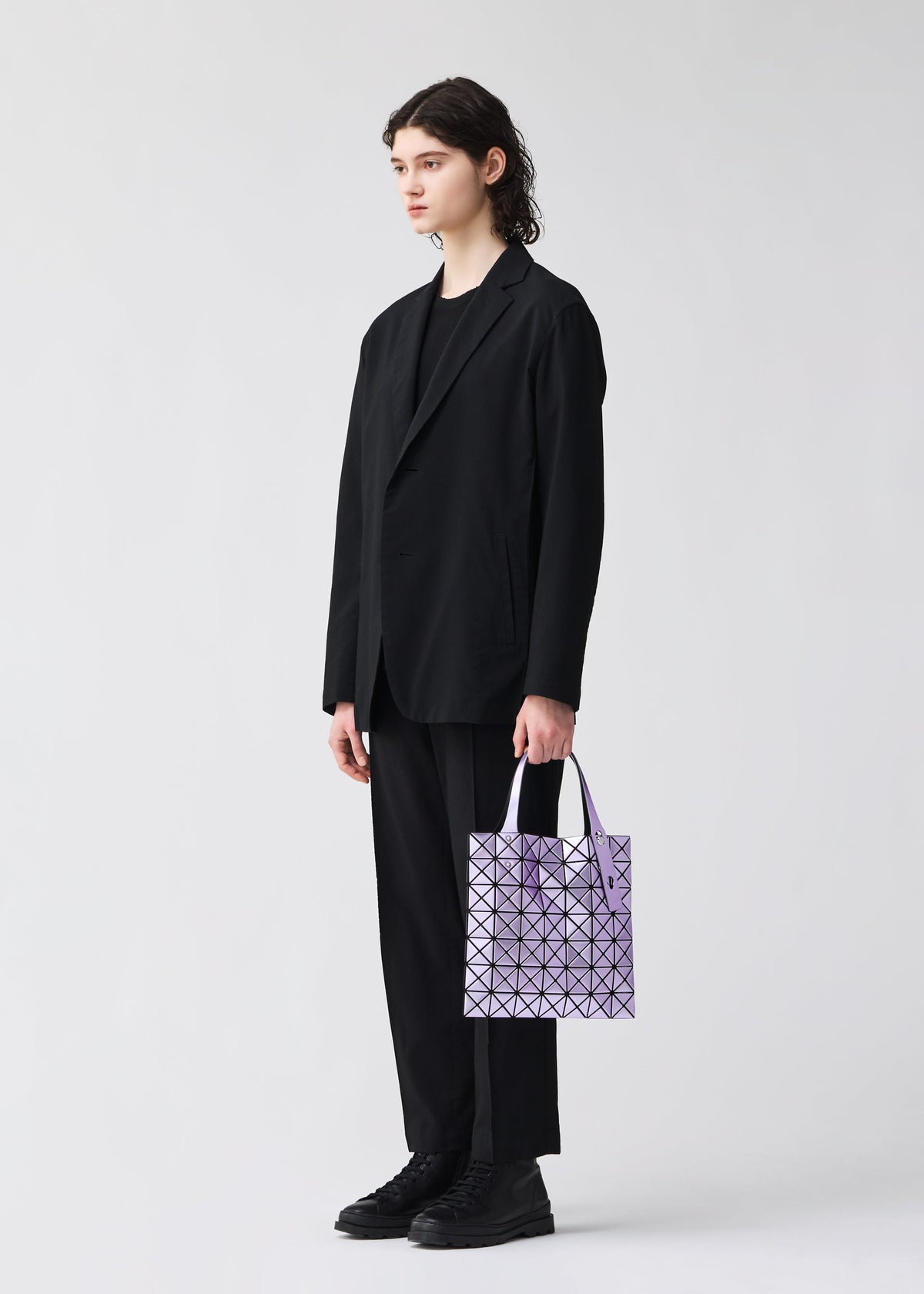 A person with shoulder-length dark hair stands against a plain light background, dressed in a black suit and shoes, holding the BAO BAO ISSEY MIYAKE PLATINUM shoulder bag with metallic triangular pieces in their left hand. view 6 of 6